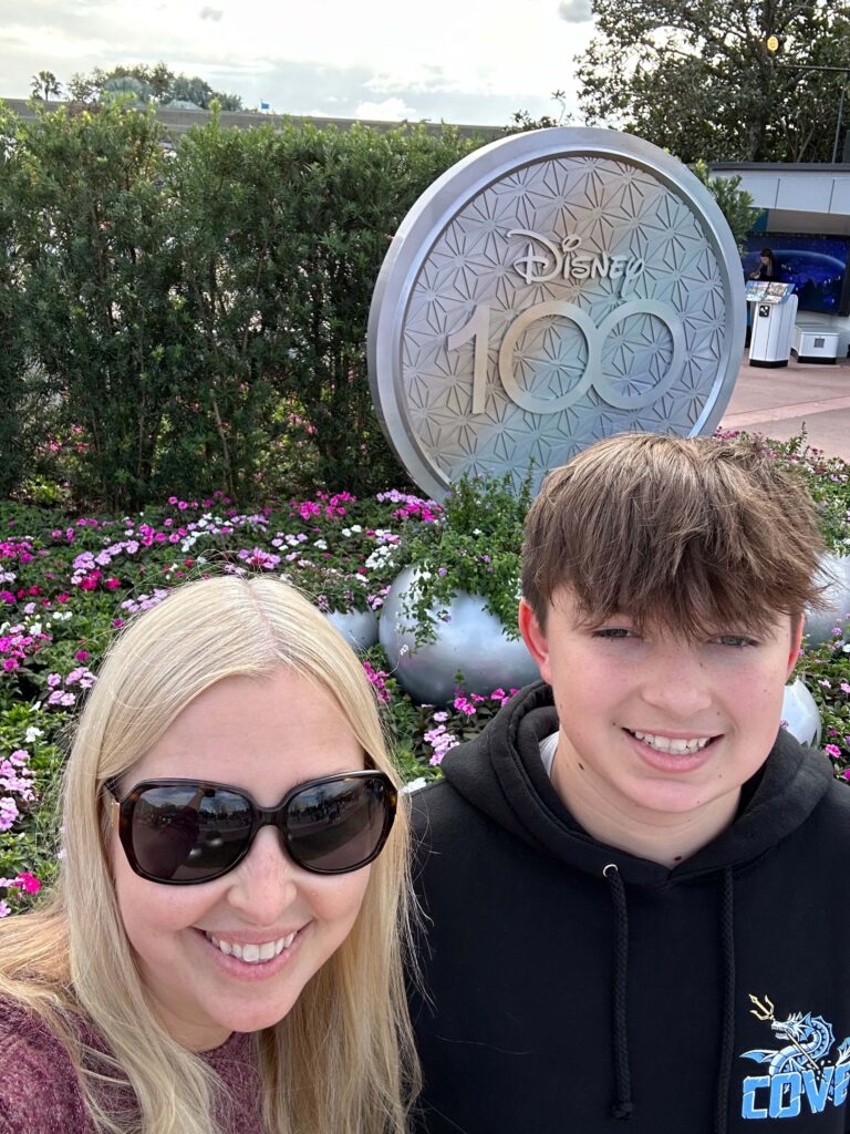 Mother and teenage son in front of the 100th anniversary Disney sign at Epcot. Seasoned Disney passholders ready to offer tips on what things to buy from Amazon for Disney.