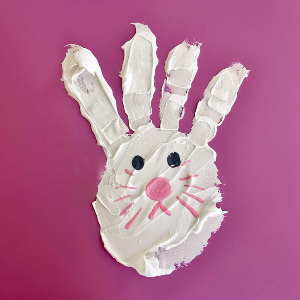 bunny handprint crafts - white paint handprint bunny, with pink and black accents for face, on a purple/pink background