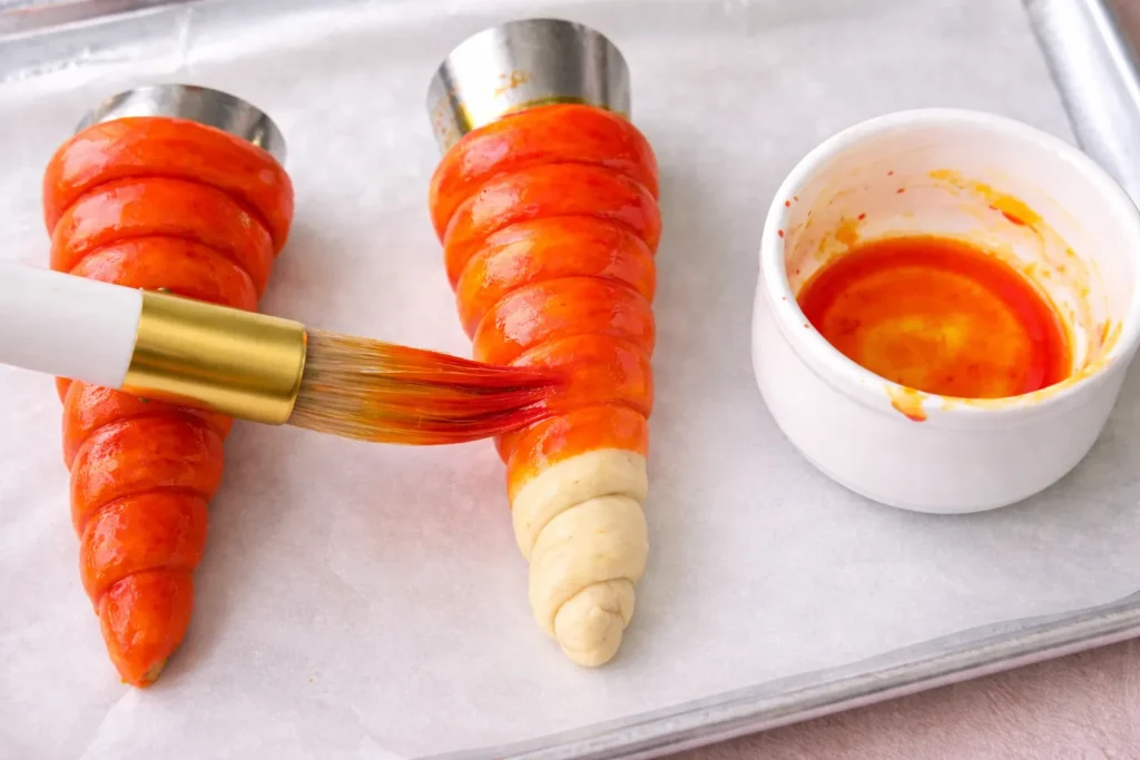 Overhead process food photography of carrot-shaped crescent dough wrapped around metal cream horn molds on a parchment lined baking sheet, one carrot being brushed with orange food coloring using a pastry brush, small white ramekin with yellow and red mixed orange coloring beside it, glossy raw dough, realistic kitchen prep scene, clean bright lighting, no text, no watermark 