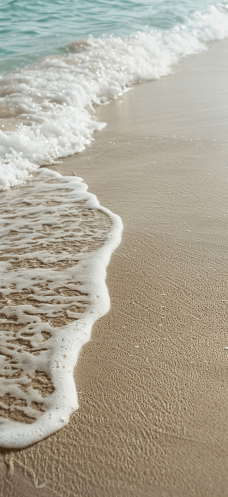 A close-up of sandy shores with gentle waves rolling in, soft beige sand and light blue water creating a calm scene.