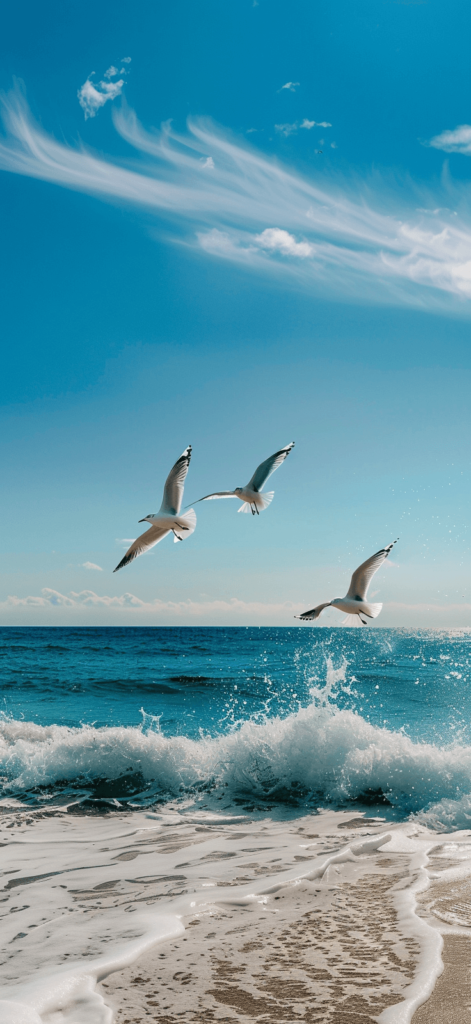 Seagulls flying over the ocean, white birds against a blue sky, with gentle waves and a sandy beach below.