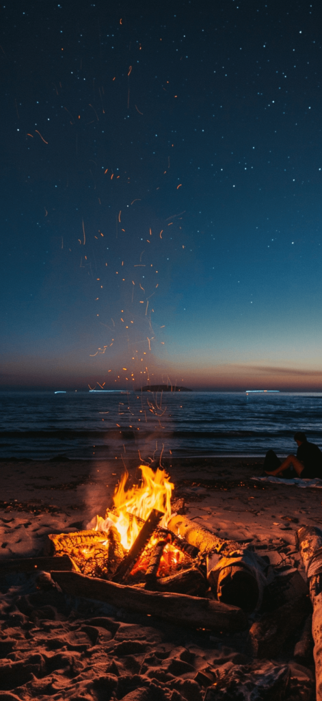 A peaceful night at the beach with a large bonfire crackling in the foreground. The flames cast a warm, golden light that illuminates the sandy beach and reflects off the gentle waves of the ocean. A group of friends is gathered around the bonfire, some sitting on logs and others on blankets, enjoying the warmth and each other’s company. The night sky is clear, revealing a canopy of twinkling stars. The overall scene is cozy and inviting, capturing the essence of a perfect summer night by the sea.