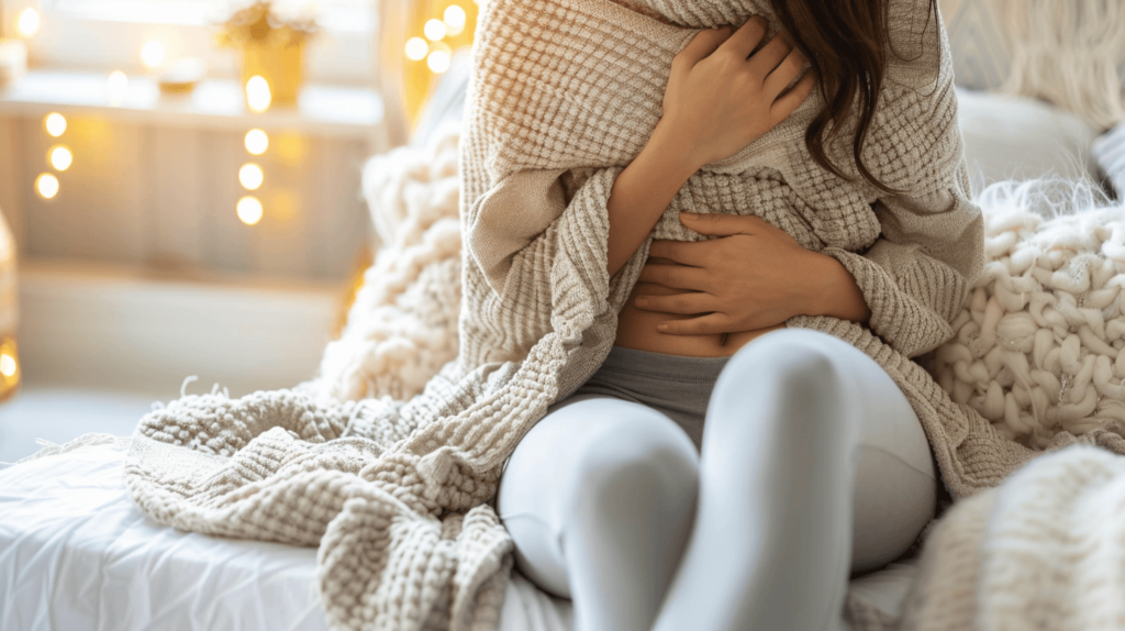 A simple image showing a woman sitting comfortably in a cozy home environment, with one hand on her stomach, indicating period cramps. She is wrapped in a soft blanket with a heating pad nearby, and the atmosphere is calm and empathetic.
