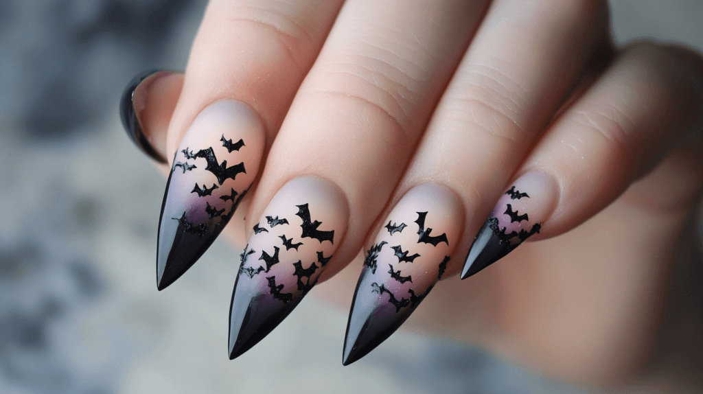 A single manicured female hand with stiletto-shaped Halloween acrylic nails, featuring a 3D design of small plastic bats attached to a black and purple ombré background.