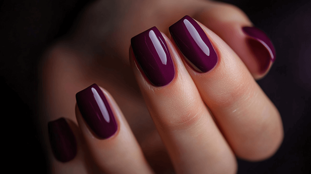 A plum purple manicure on a female hand, nails are short and square with a glossy finish.
