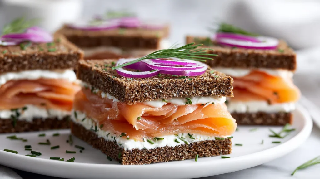 Smoked salmon tea sandwiches on thin rye bread with lemon chive cream cheese, sliced into slim fingers and arranged on a white plate in bright natural light.