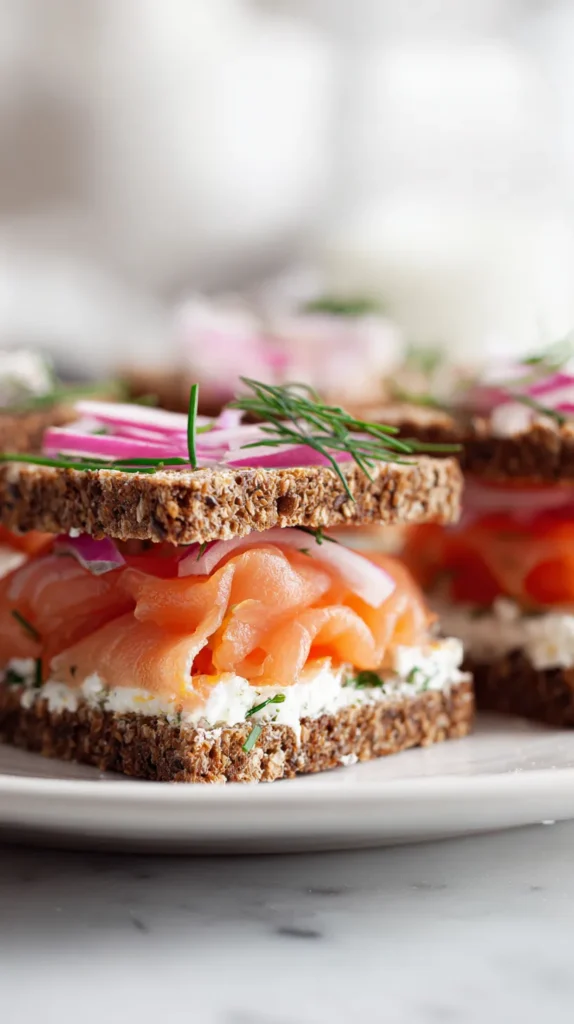 Close-up of smoked salmon tea sandwiches on dark rye with smooth cream cheese and fresh chives, cut into narrow finger shapes and styled on a white marble counter.