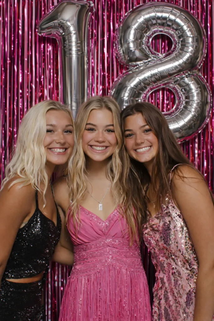 three girl friends in front of a hot pink fringe curtain background with the number 18 in silver mylar balloons