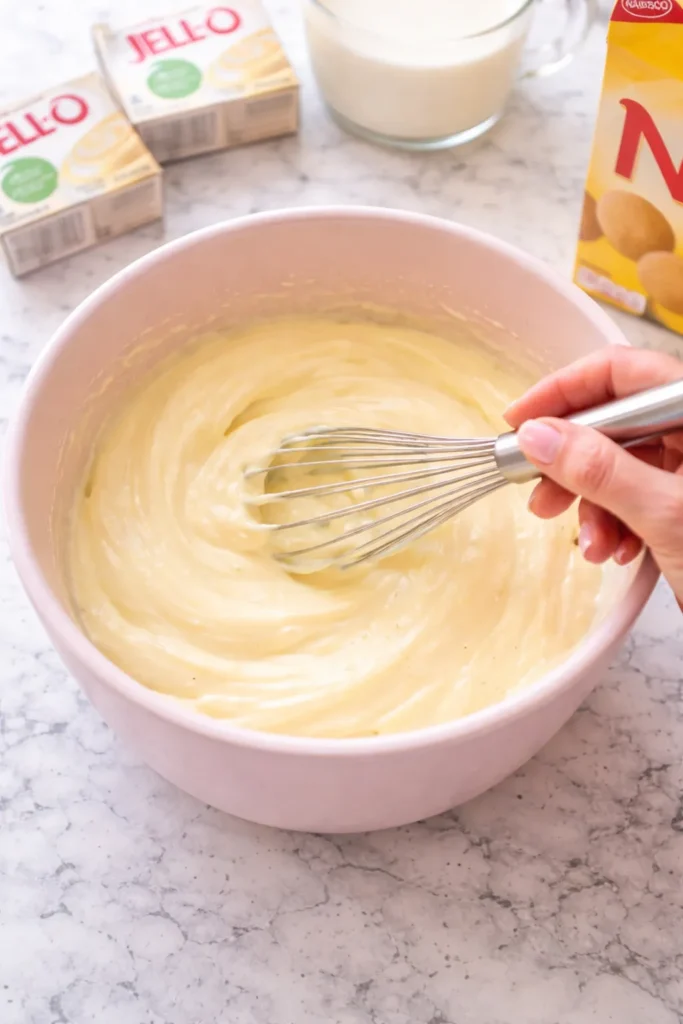 Female hand with a light pink manicure whisking vanilla pudding mix and milk in a light pink mixing bowl on a white marble countertop.