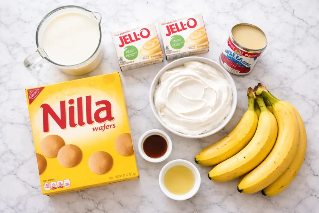 Overhead view of banana pudding ingredients on a white marble counter, including Nilla Wafers, vanilla pudding mix, sweetened condensed milk, milk, whipped topping, vanilla extract, lemon juice, and bananas.