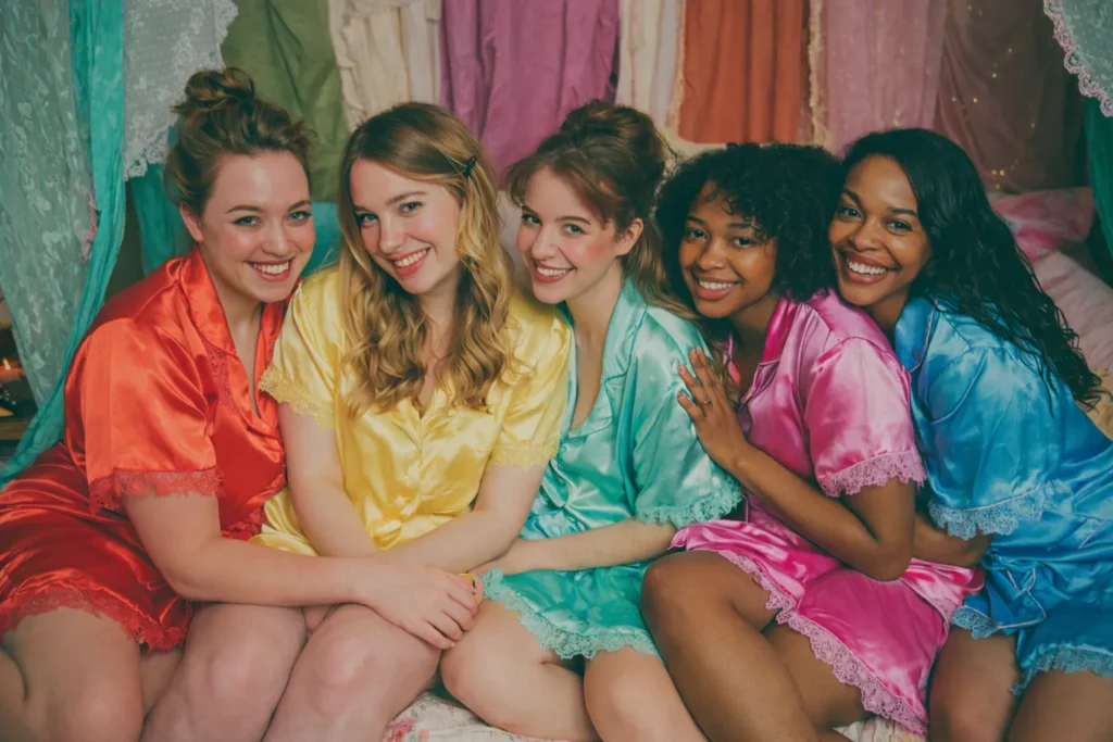 Five women in colorful satin pajamas sitting on a bed at a stylish slumber party sleepover