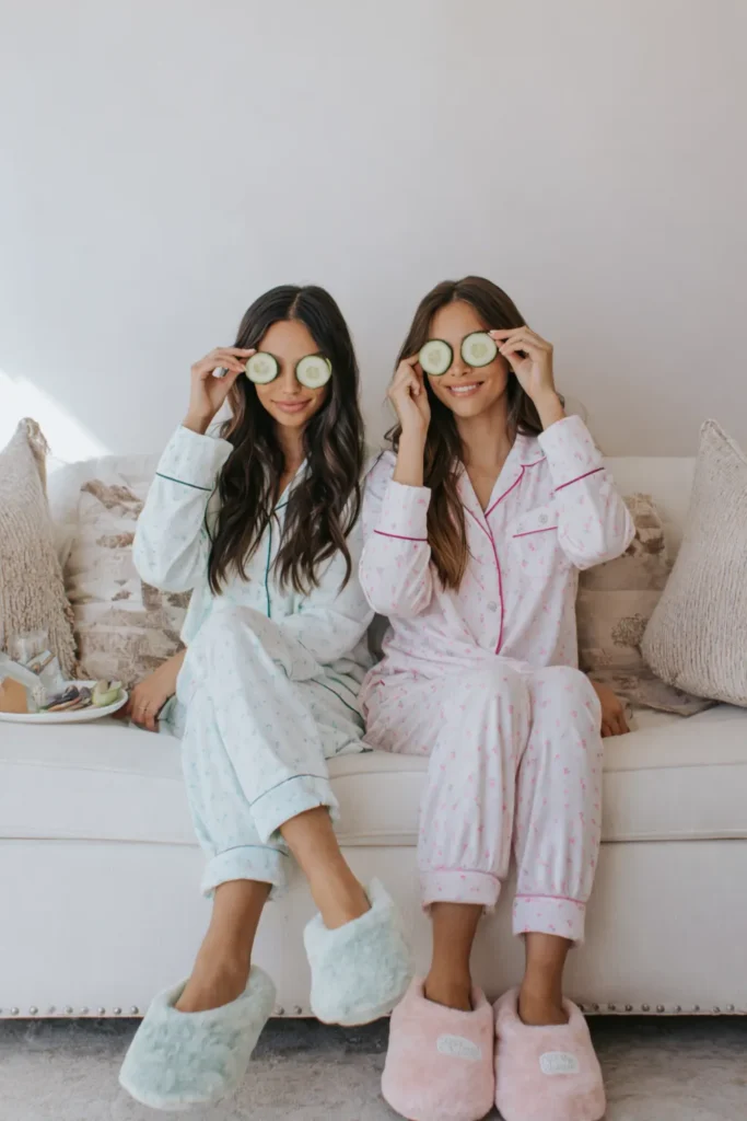 Two women in matching pajamas holding cucumber slices over their eyes on a white couch
