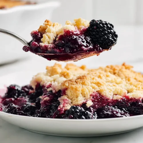 hyper-realistic photo of a serving of blackberry cobbler on a white dessert plate with a silver spoon lifting up a bite, glossy blackberry filling stretching slightly from the plate, golden cobbler topping with crisp browned edges and a soft fluffy interior, deep purple berry juices, bright white marble countertop, bright white kitchen background, soft natural window light, editorial food photography, realistic textures, no hand visible, no people, no text, no logo, no watermark