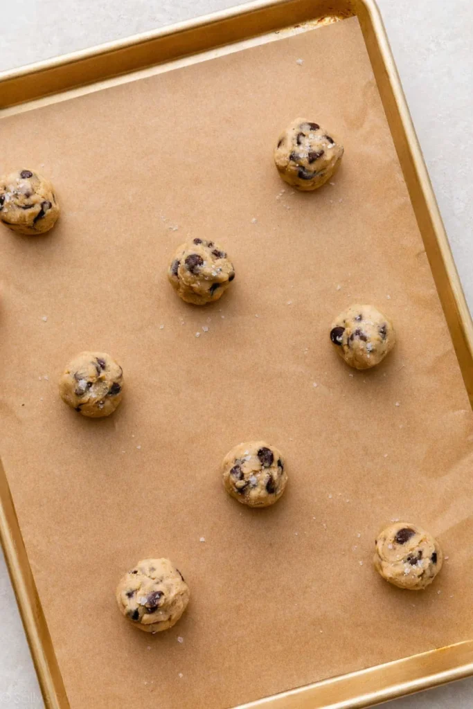 Overhead view of brown butter chocolate chip cookie dough balls on a parchment-lined gold baking sheet, topped with chocolate pieces and flaky salt.