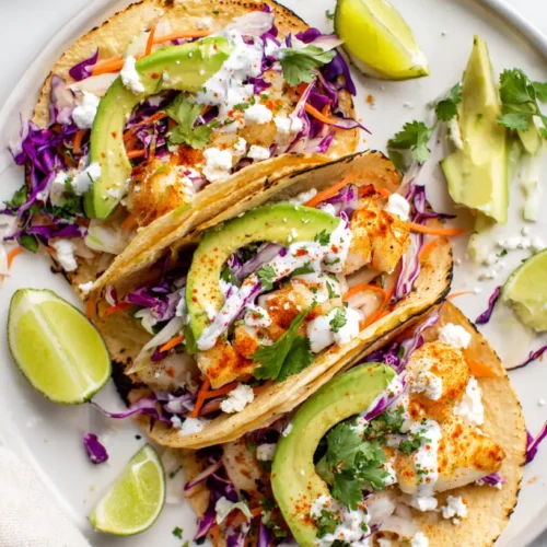 Plated healthy fish tacos filled with flaky white fish, crisp slaw, avocado, cotija, and creamy lime sauce on a white marble countertop in a bright kitchen.