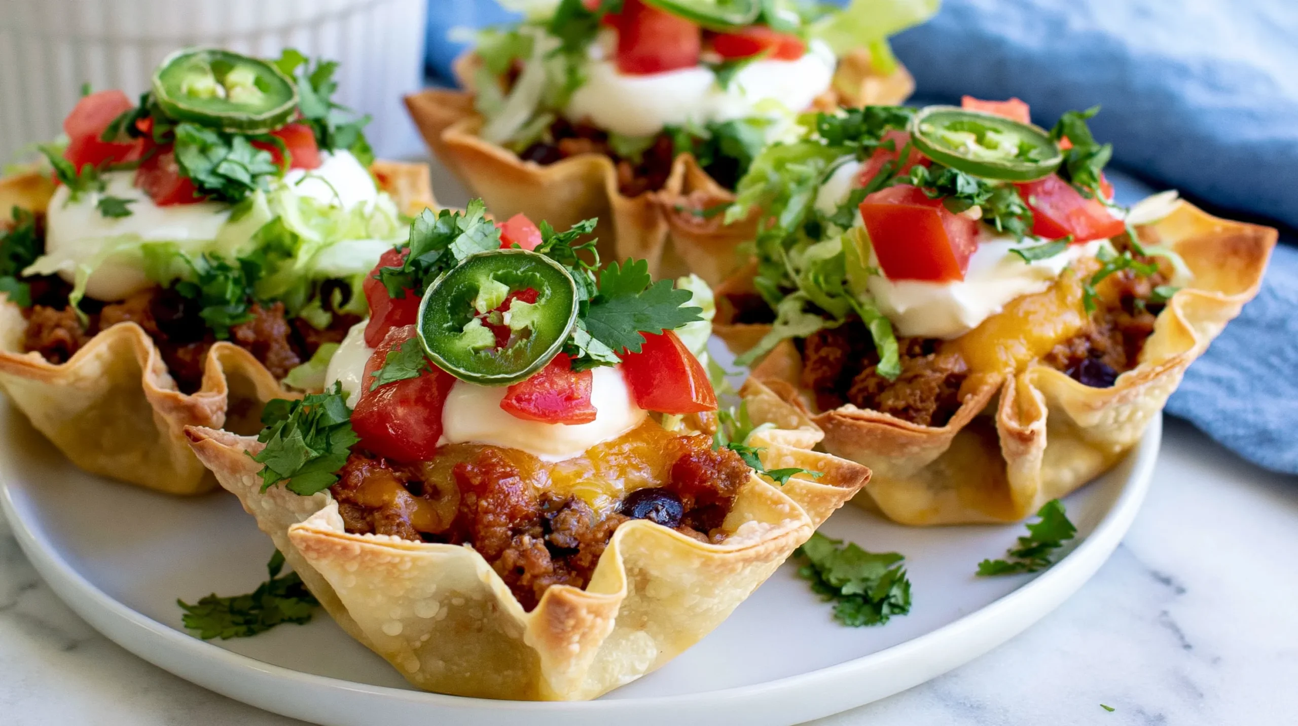 Hyper-realistic photo of taco cupcakes made with thin crispy square wonton wrappers folded into small angular cups, visible pointed wonton corners, delicate blistered golden wrapper texture, crisp baked edges, filled with seasoned ground beef, black beans, melted cheddar cheese and Monterey Jack cheese, topped lightly with sour cream, diced tomatoes, shredded lettuce, chopped cilantro, and thin jalapeño slices, small hand-held appetizer size, arranged on a simple white plate on a white marble countertop in a bright white kitchen, soft natural window light, clean editorial food photography, realistic texture, fresh colorful toppings, true-to-life proportions, slightly overhead angle, shallow depth of field, no utensils, no hands, no text, no watermark, no muffin tin, no metal baking pan, no tortilla bowls, no fluted pastry shells, no ruffled pie crust, no oversized taco salad bowls