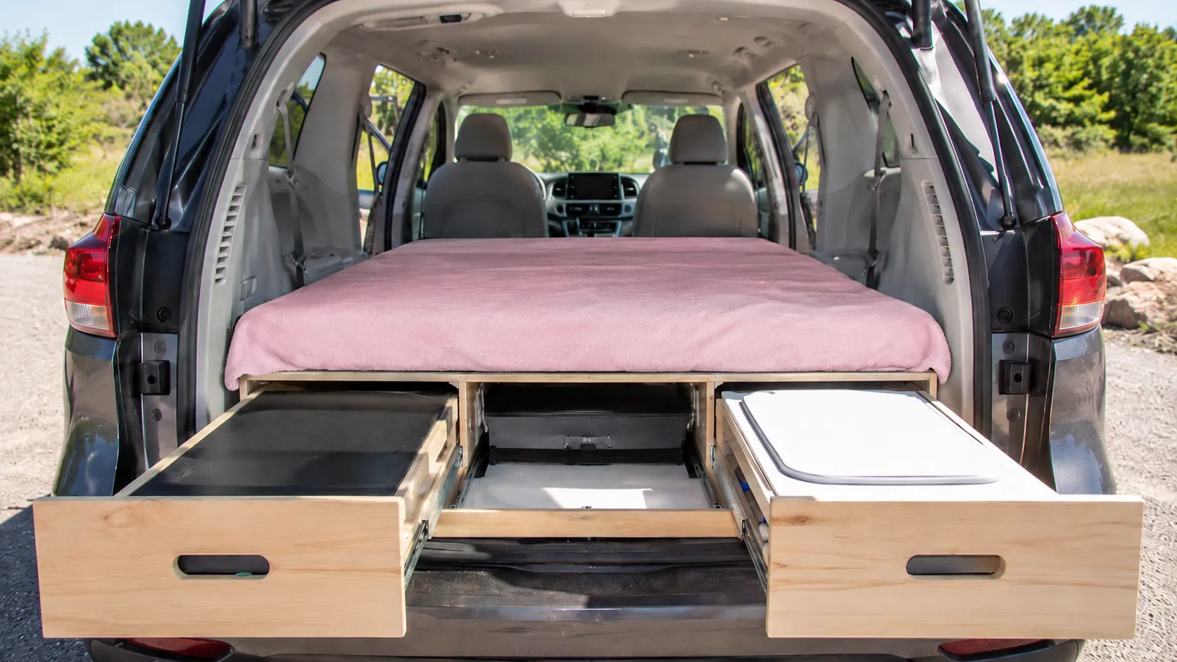 Toyota Sienna camper conversion with a pink blanket over the mattress, built-in wooden pull-out drawers, and open rear hatch.