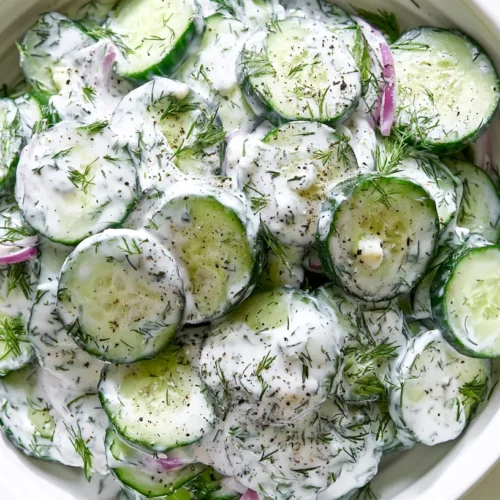 Overhead editorial food photography of cucumber dill Greek yogurt salad in a shallow white ceramic bowl on a white marble kitchen counter, made with thinly sliced unpeeled thin English cucumbers with visible dark green edges, thick plain Greek yogurt dressing, fresh lemon juice, olive oil, finely grated garlic, fresh chopped dill, fresh chopped parsley, thin slices of red onion throughout the salad, cracked black pepper, and a light glossy coating from the dressing, cucumber slices cut very thin and evenly, creamy dressing clearly clinging to the cucumbers, fresh cold summer salad, soft natural window light, bright white kitchen background, clean fresh styling, realistic textures, no text, no watermark