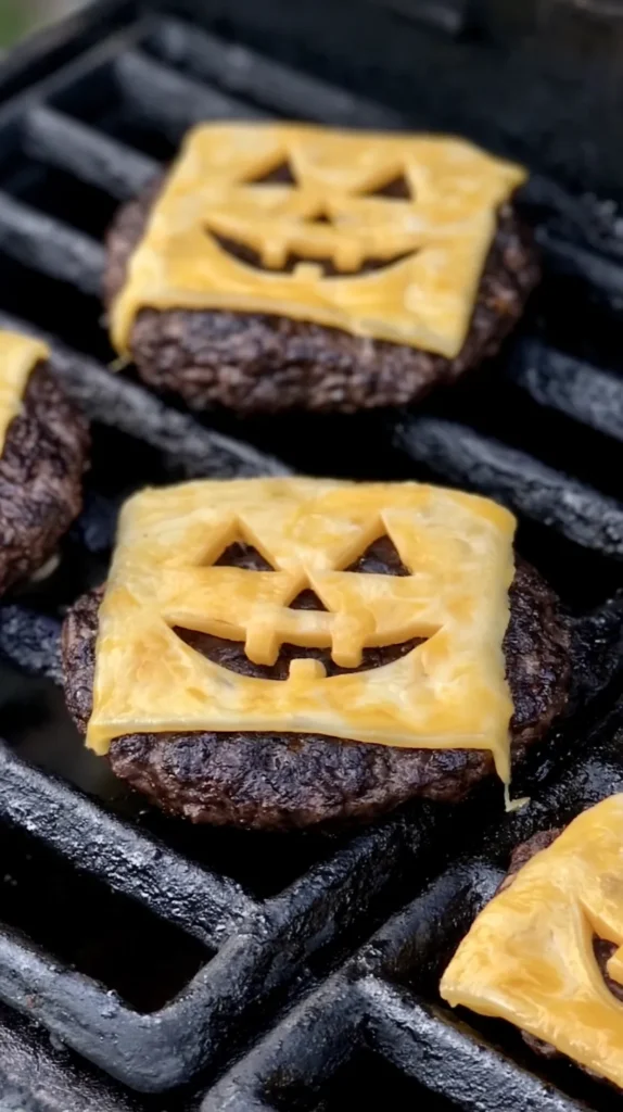 a close up shot of three unique burgers on a grill, each featuring a slice of cheese cut into a jack o' lantern design. the cheese is partially melted, cheese slightly melting over the burger sides. the burger patties are well cooked and dark brown. the grill is robust with black, thick bars, some of which show wear and tear. the items are arranged in a triangular formation, occupying the central and lower right portions of the frame.