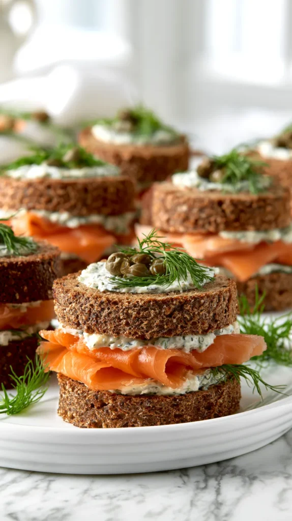 hyper-realistic editorial food photography of smoked salmon tea sandwiches, soft rye and pumpernickel sandwich bread with crusts removed, cut into small neat closed rounds with a top and bottom bread layer, creamy herb spread visible between the bread, thin folded smoked salmon layered inside, tiny capers and fresh dill peeking from the edges, clean polished cut edges, arranged neatly on a white ceramic platter, white marble kitchen counter, bright white kitchen background, soft natural window light, elegant afternoon tea styling, realistic textures, no people, no hands, no text, no watermark