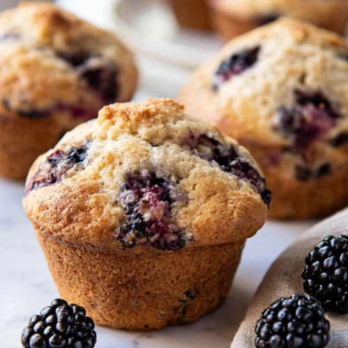 Hyper-realistic photo of freshly baked blackberry muffins on a white marble kitchen counter, soft golden domed tops, juicy dark purple blackberry pockets visible on the surface, a few fresh blackberries scattered nearby, neutral linen napkin, bright natural window light, clean summer kitchen background softly blurred, cozy homemade bakery feel, realistic muffin texture, no text, no watermark, no people