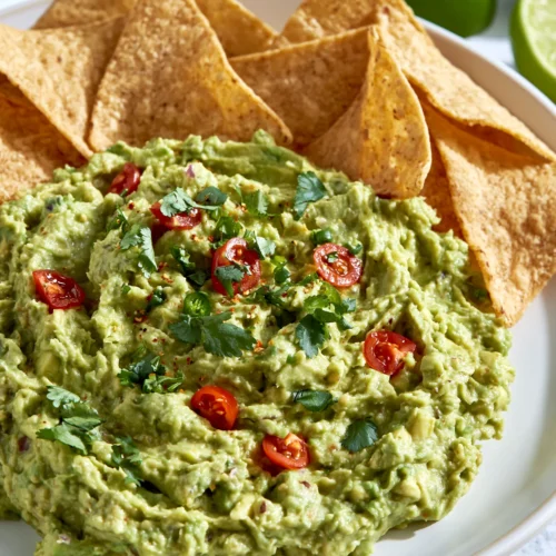 Fresh guacamole dip topped with red chili slices and cilantro, served with crispy tortilla chips and lime wedges on a white plate