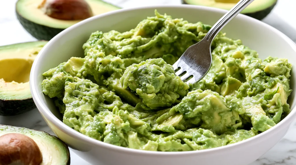 White bowl filled with creamy green guacamole and a fork, surrounded by fresh avocado halves on marble surface