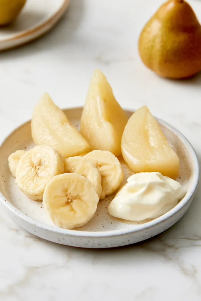 Sliced pears and banana pieces with a dollop of yogurt on a speckled white bowl