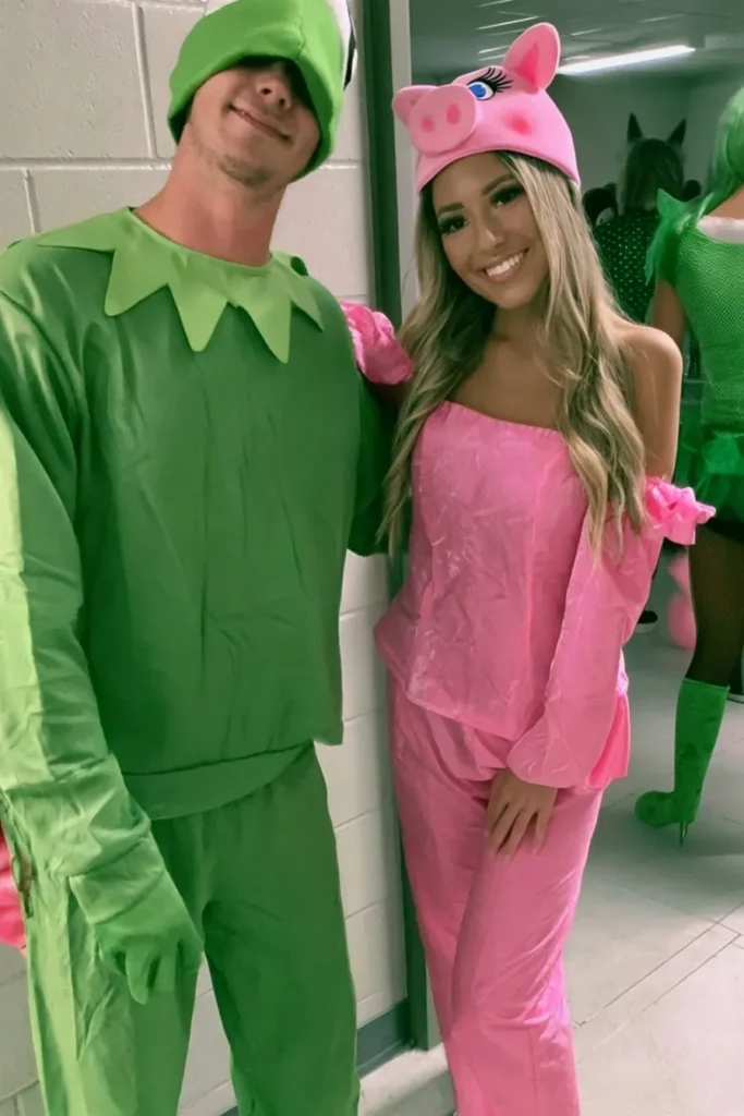 Couple in matching Muppet costumes—man dressed as Kermit the Frog in green with lily pad collar and woman as Miss Piggy in pink with pig ears headband, posing in a bathroom mirror