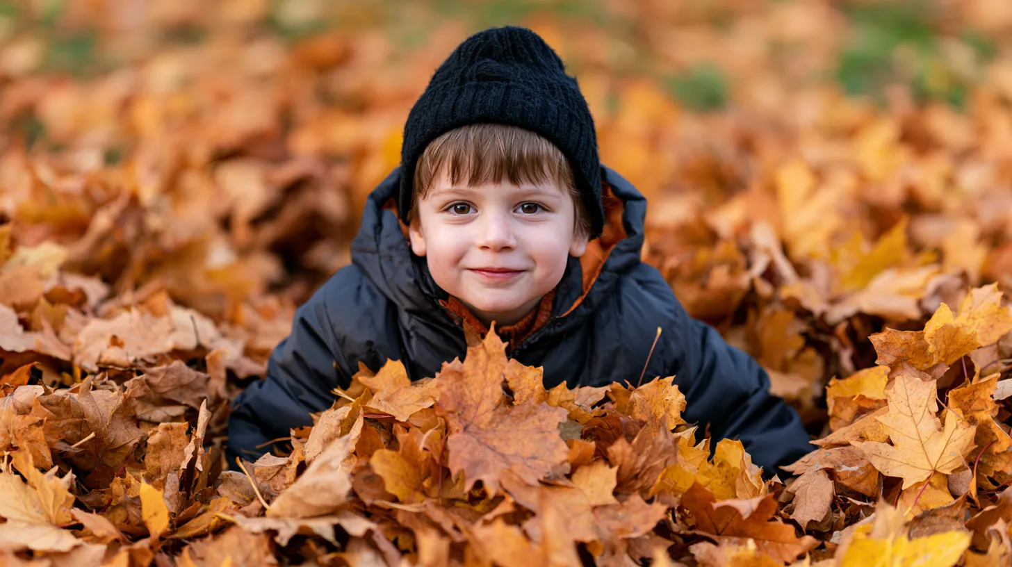 Smiling young boy in black jacket and beanie lying in a pile of golden autumn leaves