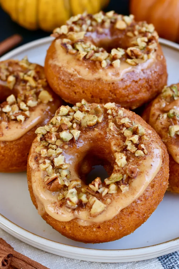 Glazed donuts topped with crushed pecans and cinnamon sugar on a white plate with pumpkins in the background
